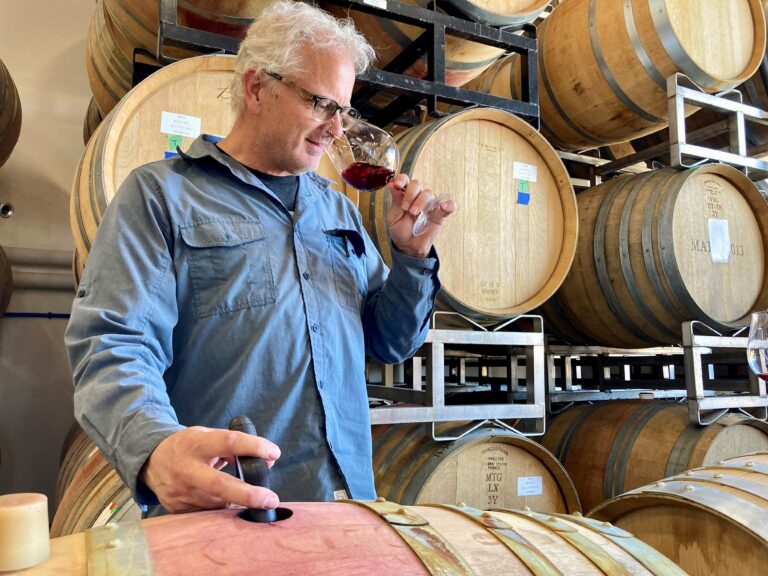 man standing, surrounded by oak barrels, tasting a glass of red wine