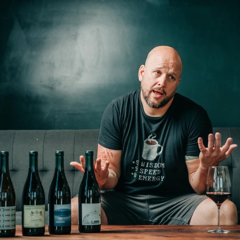 Man gesturing with hands while he speaks, sitting next to a line up of wine bottles
